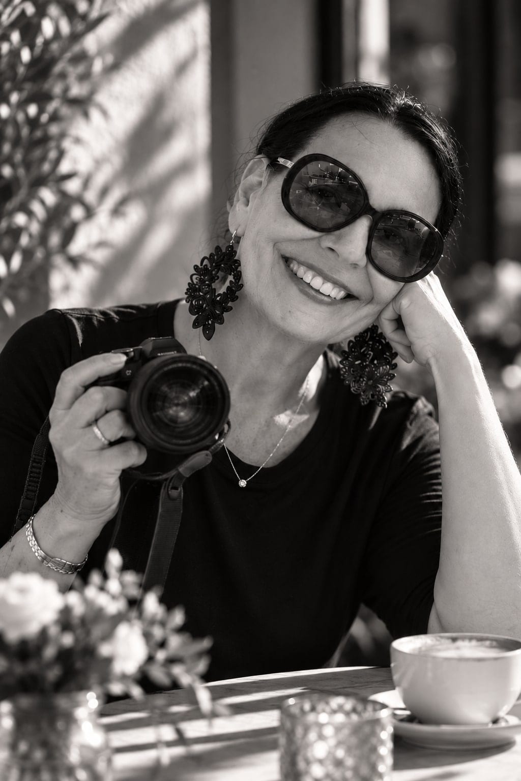 Kerstin Klesse an einem Cafétisch, lachend, mit Sonnenbrille und Kamera in der Hand, Schwarz-Weiß
